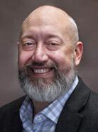 A bald man with a gray beard, wearing a checkered shirt and dark blazer, smiles against a plain, dark background.