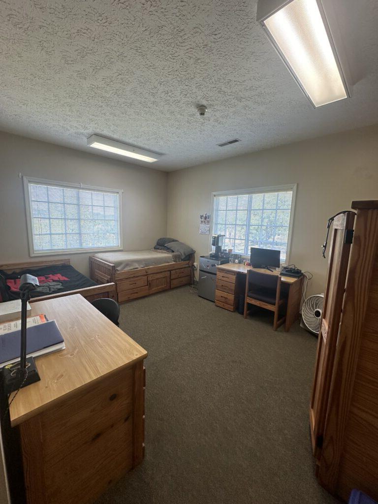 A college dorm room with a bed, desk, dresser, refrigerator, and computer setup. Two windows let in natural light.