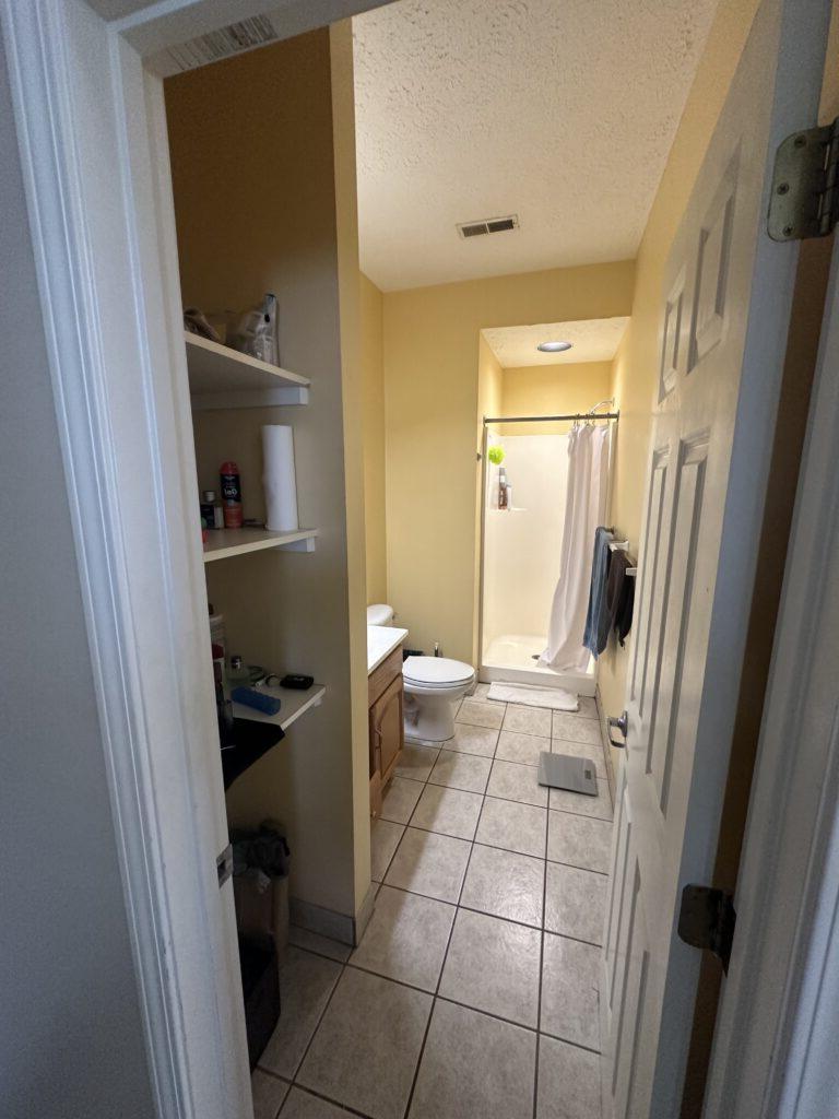 A small bathroom with a shower, toilet, and sink. Open shelves on the right contain various items. The walls are yellow, and the floor has white tiles. A white door is partially open.