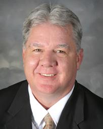 A man with short gray hair, wearing a dark suit and a light-colored tie, smiles at the camera against a neutral background.